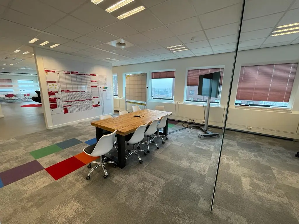 Vergaderruimte op het Johanna Westerdijkplein met een houten tafel, witte stoelen, grote ramen en een overzichtsbord aan de muur.